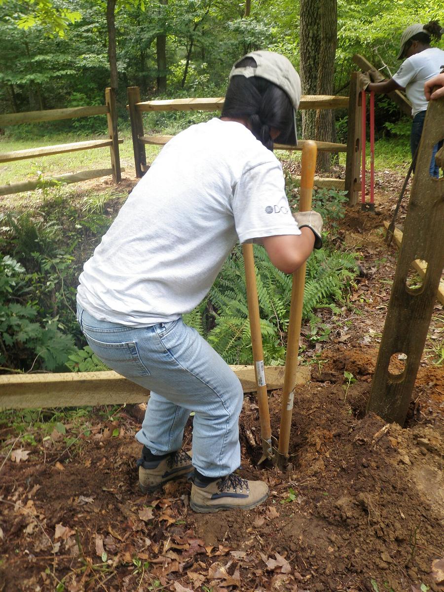 A worker digging a post hole with a manual post hole digger beside a wooden fence